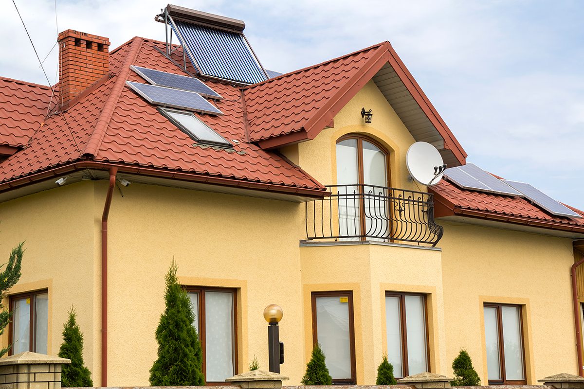 Close-up of new brick house top with red shingle roof, satellite dish, solar panels and plastic attic windows on bright blue sky background.